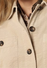 Beżowa kurtka z kordurowej tkaniny o teksturowej powierzchni, z dwoma dużymi brązowymi guzikami i kieszenią na piersi. Warstwowa, z wzorzystą koszulą pod spodem.