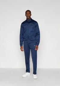 Tumši zils sporta tērps no gludas auduma, ar rāvējslēdzēju jaku un biksēm ar elastīgu vidukli, kopā ar baltām sporta kurpēm.