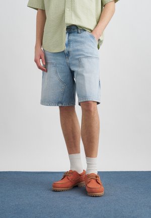 Άτομο που φοράει ανοιχτό μπλε τζιν σορτς, άσπρες κάλτσες και πορτοκαλί παπούτσια με κορδόνια, στέκεται σε μπλε χαλί με το ένα χέρι στην τσέπη.