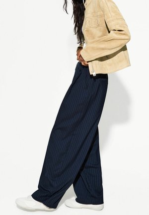 Beige mockajacka med präglad "FUEL"-logga, kombinerad med marinblå vida byxor med vertikala pinstripes och vita sneakers.