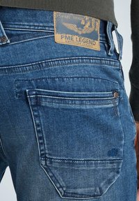 Jeans de mezclilla azul con un acabado desgastado. Presentan un parche de cuero en la cintura y bolsillos traseros cosidos. Acentos de remaches y botones visibles.