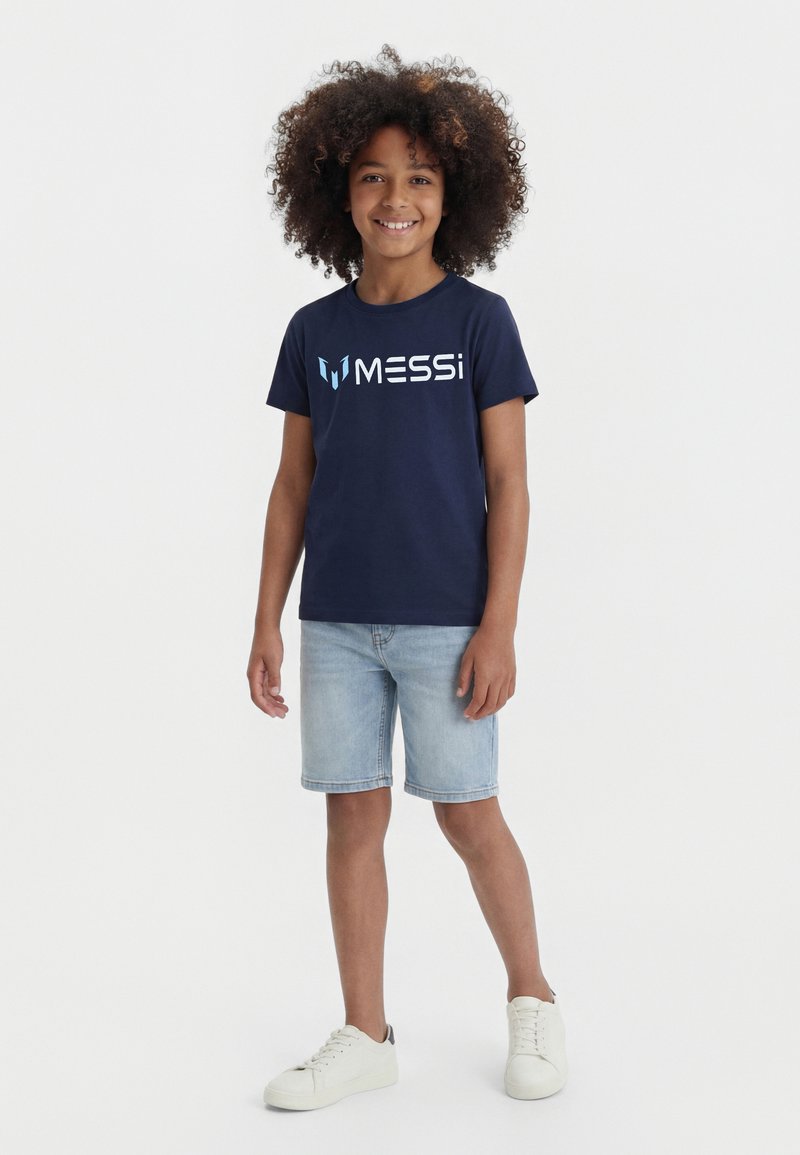Lachend kind met krullend haar, gekleed in een marineblauw "Messi" t-shirt, lichte spijkershorts en witte sneakers, staand tegen een effen witte achtergrond.