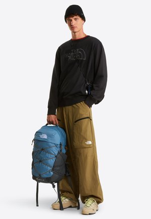 Uomo con berretto nero, felpa nera The North Face, pantaloni kaki e scarpe beige che tiene uno zaino The North Face blu e nero.