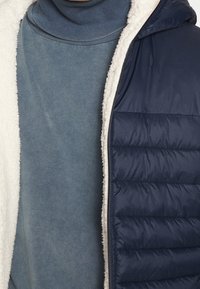 Marinblå vadderad väst med vit fleecefoder, med kviltade sektioner. Under, en ljusblå polotröja i sweatshirtmaterial. Mjuk textur.