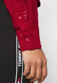 Red corduroy shirt cuff with two round buttons and stitching details. Black pants with white "TOMMY JEANS" logo on the side.