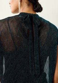 Prozirna crna bluza s teksturiranim uzorkom i mašnom za zatvaranje straga. Tkanina suptilno sjaji, dodajući dubinu dizajnu.
