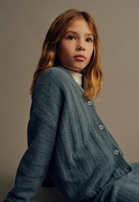 Jopica iz mehkega, teksturiranega modrega blaga z rebrastim vzorcem, okroglim izrezom in velikimi gumbi, nošena čez belo srajco.