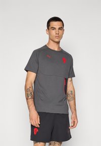 Grå sports t-shirt med et rødt Puma-logo, kontrasterende rød lynlås-lomme og korte ærmer, parret med mørke shorts med et rødt design.