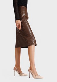Gonna a matita in ecopelle marrone con spacco laterale, abbinata a décolleté nude con punta affusolata e dettaglio decorativo a fibbia.