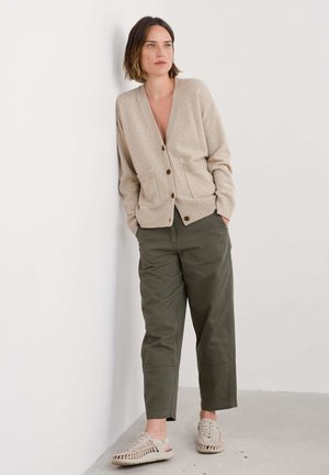 Cardigan beige con scollo a V, polsini a costine e tasche frontali. Pantaloni a palazzo verdi oliva. Sneakers beige con texture e dettagli intrecciati.