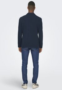 Marineblazer mit strukturierten Schultern, mit einem Schlitz hinten, kombiniert mit eng anliegenden blauen Jeans und weißen Low-Top-Sneakern.
