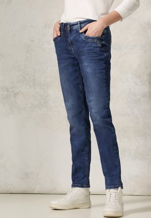 Jeans in denim blu con vestibilità slim, design a cinque tasche e cuciture sottili. Indossati con sneakers bianche alte.