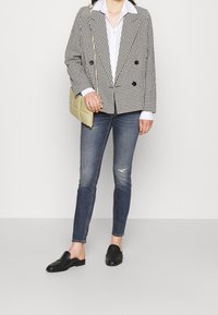 Blazer de cuadros en blanco y negro de doble botonadura, camisa blanca, jeans azul desgastados y zapatos de cuero negros tipo slip-on, con un bolso de hombro beige.
