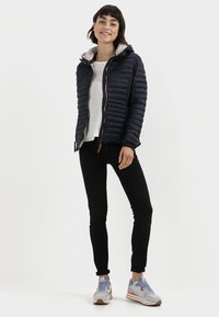 Navy puffer jacket with a hood, quilted texture, front zipper, and tan accents. Paired with black pants and light blue athletic shoes.