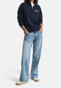 Persoon draagt een marineblauwe trui met kwartzip en wit logo, lichtblauwe jeans met wijde pijpen en beige sneakers, staat met handen in de zakken.