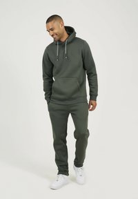 Sweat à capuche vert foncé avec une poche kangourou, des cordons de serrage, et un pantalon de jogging assorti ; le tissu semble doux avec une texture lisse ; accompagné de baskets blanches.