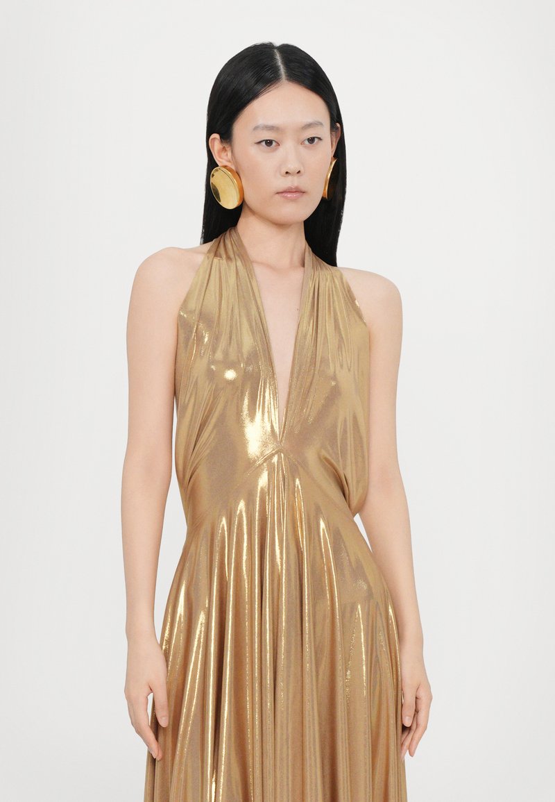 Abito halter dorato con scollatura a V profonda, caratterizzato da una texture metallica lucente e una silhouette fluida, completato da grandi orecchini circolari.