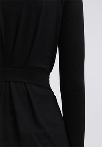 Černé šaty s dlouhým rukávem, které mají přiléhavý pas a nabíranou sukni, vyrobené z hladkého materiálu, s jednoduchým designem a bez viditelných vzorů.