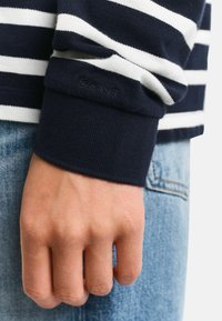 Marineblauw en witte gestreepte lange mouwen shirtmanchet met ribbeltextuur en geborduurd "GANT" logo. Hand die denim jeans vasthoudt.