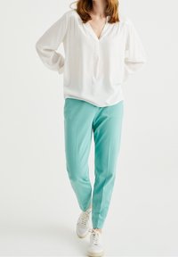 Vit långärmad blus med V-ringning och subtil textur, kombinerad med ljus turkos byxor och vita sneakers med texturerade accenter.