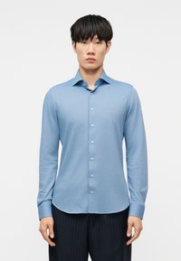 Νέος άνδρας που φορά ανοιχτό μπλε πουκάμισο με κουμπιά μακριάς μανίκας και σκούρο παντελόνι με λεπτές λωρίδες, στέκεται μπροστά σε απλό λευκό φόντο.