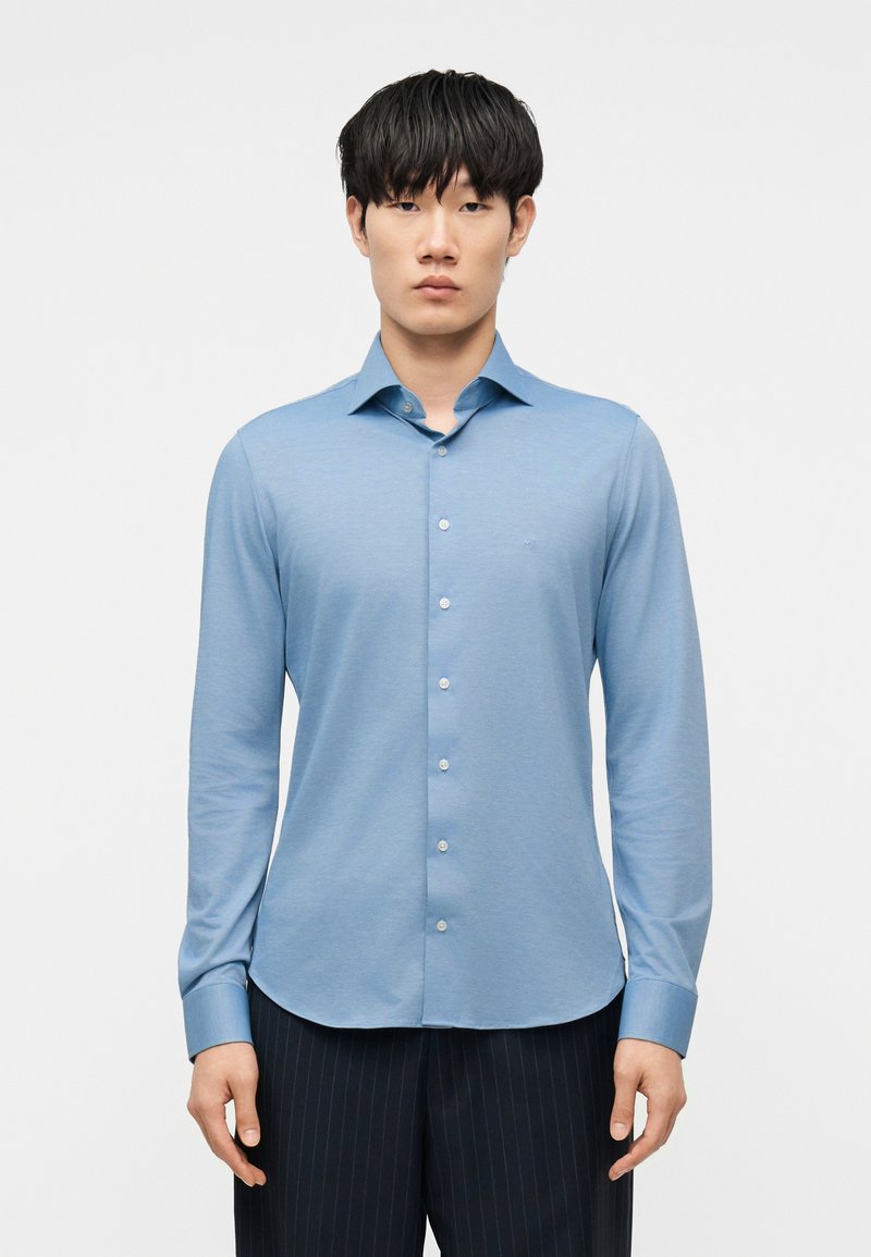 Νέος άνδρας που φορά ανοιχτό μπλε πουκάμισο με κου μπιά μακριάς μανίκας και σκούρο παντελόνι με λεπτές λωρίδες, στέκεται μπροστά σε απλό λευκό φόντο.