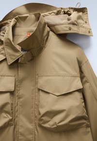 Μπεζ αδιάβροχο μπουφάν με γιακά που στέκεται, εσωτερική επένδυση από δίχτυ, δύο τσέπες στο στήθος και κούμπωμα με κουμπιά στο μπροστινό μέρος. Διαθέτει κουκούλα με κορδόνι.