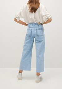 Femme aux longs cheveux portant un chemisier blanc, un jean large court bleu clair, et des baskets blanches, debout les mains sur les hanches, vue de dos.