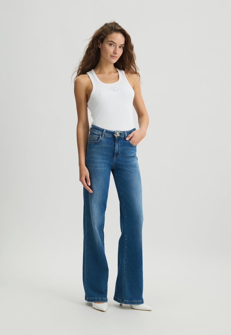 Weißes Ripp-Top mit eingeprägtem Logo, kombiniert mit hochgeschnittenen blauen Jeans mit einer dezenten Ausbleichung und weit geschnittenem Design.