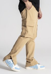 Beżowe spodnie cargo z wieloma kieszeniami, prosta nogawka i gładka faktura, w zestawieniu z niebieskimi i białymi trampkami w wzory.