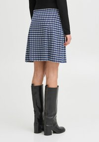 Jupe en tartan bleu avec des plis, présentant une texture douce, assortie à des bottes en cuir noir montantes avec un large talon et des coutures contrastées.