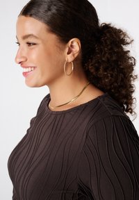 Jonge vrouw die glimlacht, met een gestructureerde donkere top, gouden hoepel oorbellen, een gouden ketting, en krullend haar dat in een paardenstaart is vastgebonden.