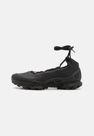 Zwarte instapschoenen van synthetisch materiaal, met een rubberen zool met textuur voor grip, een ronde neus en verstelbare veters bovenaan.