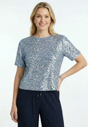 Femme souriante, portant un haut à paillettes argenté brillant à manches courtes et un pantalon plissé bleu foncé, une main sur la hanche sur fond blanc.