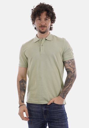 Uomo con capelli ricci e tatuaggi su entrambe le braccia, indossa una polo verde chiaro e jeans blu, in piedi con una mano in tasca.