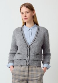Grijze gebreide cardigan met een V-hals, contrasterende stiksels en zilveren knopen. Bevat twee zakken en wordt gedragen over een lichtblauw overhemd met kraag.