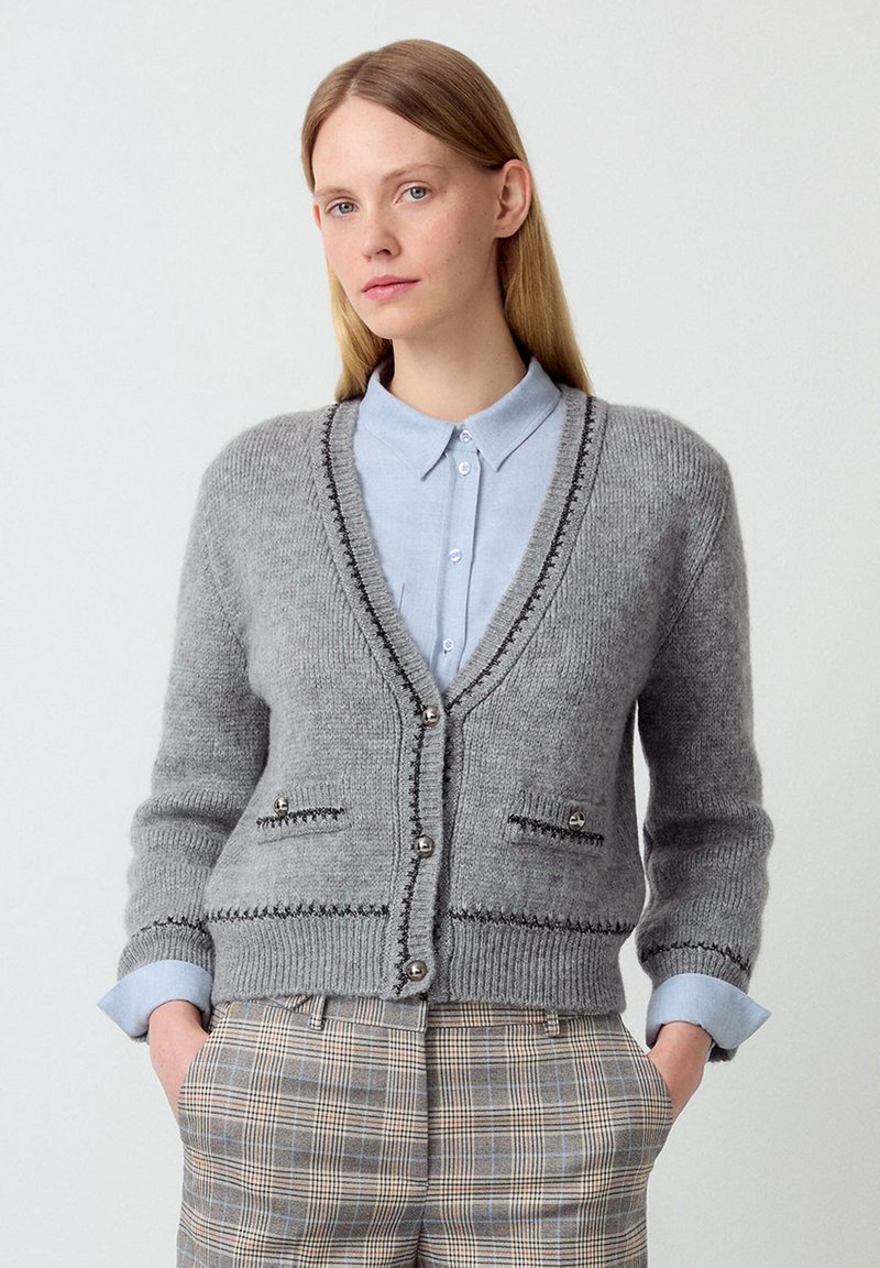 Grauer Strickcardigan mit V-Ausschnitt, kontrastierenden Nähten und silbernen Knöpfen. Verfügt über zwei Taschen und wird über einem hellblauen Hemd mit Kragen getragen.
