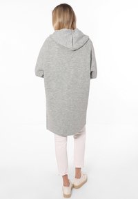 Felpa grigia oversize con cappuccio, caratterizzata da una texture morbida, orlo arrotondato e vestibilità comoda, abbinata a pantaloni chiari e sandali casual.