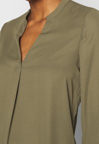 Oliwkowa bluzka uszyta z gładkiego materiału, z dekoltem w kształcie litery V i delikatnymi plisami z przodu. Długie rękawy z subtelnym przeszyciem.