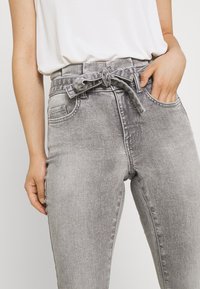 Jeans de mezclilla gris con cintura alta, cinturón de lazo, dos bolsillos frontales y una textura ligeramente desgastada. Modelo vistiendo una blusa blanca.