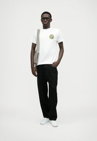 Νέος άνδρας που φοράει γυαλιά, λευκό graphic t-shirt, μαύρο παντελόνι, λευκά αθλητικά παπούτσια και γκρι τσάντα ώμου, στέκεται μπροστά σε ένα απλό λευκό φόντο.