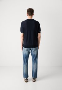 Mornarsko polo majica s kratkimi rokavi, z belimi črtastimi poudarki na ovratniku in rokavih, v kombinaciji s svetlo modrimi jeans hlačami ravnega kroja in športnimi čevlji.