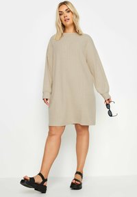 Abito beige a coste con maniche lunghe e scollatura rotonda. La modella indossa sandali con plateau neri e tiene occhiali da sole neri in una mano.