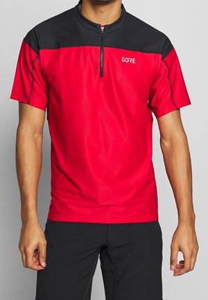 Mann trägt kurzärmeliges rotes und schwarzes Sportshirt mit Reißverschluss und "GORE"-Logo, kombiniert mit schwarzen Shorts, steht vor einfachem Hintergrund.