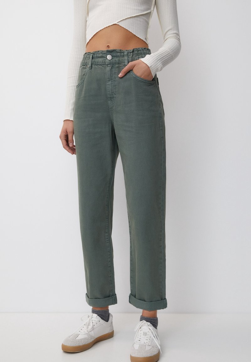 Personne portant un pantalon vert taille haute retroussé avec des poches, des baskets blanches, des chaussettes grises et un haut blanc court à manches longues.