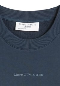 Μπλε ναυτικό πουλόβερ με λευκή ετικέτα και αντίστοιχο κείμενο στο στήθος που γράφει "Marc O'Polo DENIM" σε ανοιχτό μπλε και λευκό.