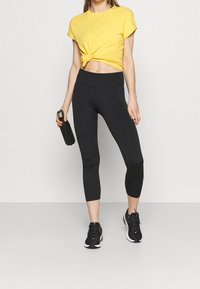 Top giallo a maniche corte con nodo frontale, leggings neri a mezza gamba e scarpe da ginnastica nere, mentre tiene una borraccia scura.