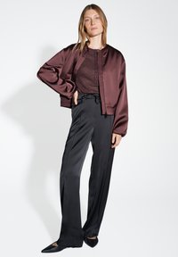 Giacca bomber in satin bordeaux, pantaloni neri a gamba larga, top strutturato; design elegante, tessuti lisci e scarpe nere a punta.