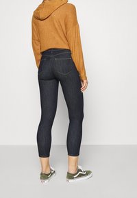 Jeans skinny de un índigo oscuro con una cintura alta, combinados con un suéter marrón con capucha y zapatillas verdes con suelas blancas.