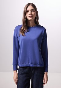 Blauwe langwerpige sweatshirt met een ronde hals, geribde zoom en een relaxte pasvorm. Gladde textuur, gecombineerd met een donkere spijkerbroek.